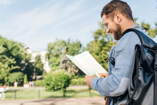 Seitenansicht des jungen lächelnden tragenden Rucksacks des Mannes und der Lesekarte an draußen