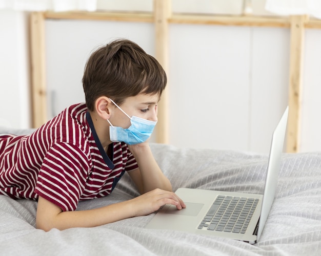 Seitenansicht des Kindes mit der medizinischen Maske, die auf Laptop spielt