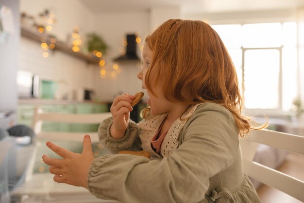 Seitenansicht des kleinen hellhäutigen rothaarigen Mädchens, das mit Vergnügen Kekse isst, während es zu Hause am Tisch sitzt Konzept für Essen und Kinder