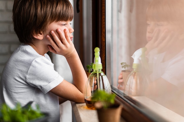 Kostenloses Foto seitenansicht des kleinen jungen, der durch das fenster mit wasserspray für ernten schaut