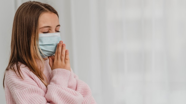 Kostenloses Foto seitenansicht des kleinen mädchens, das mit medizinischer maske und kopienraum betet