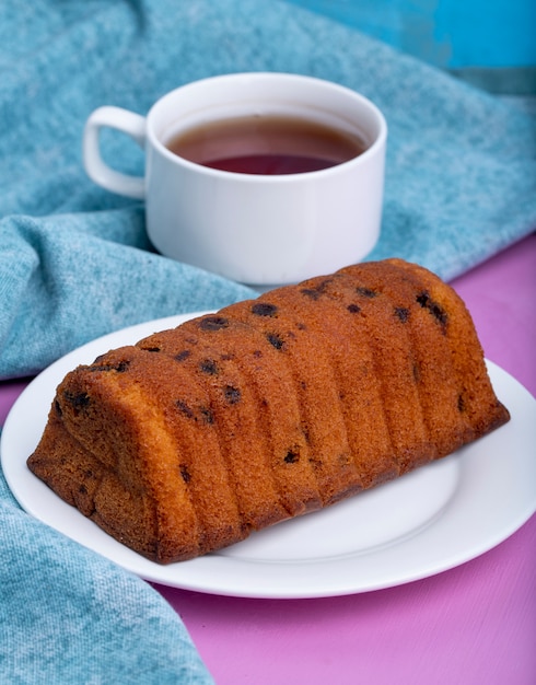 Seitenansicht des Kuchens mit Rosinen auf einem weißen Teller und einer Tasse schwarzen Tees auf lila und blau