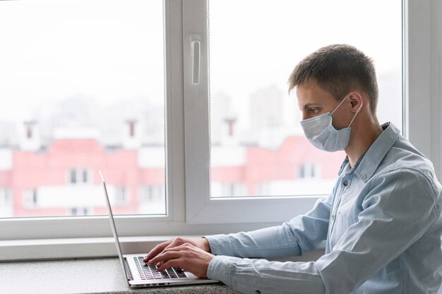 Seitenansicht des Mannes mit der medizinischen Maske, die am Laptop arbeitet