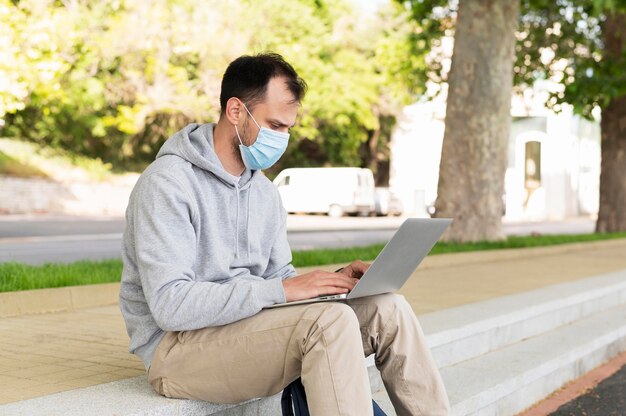 Seitenansicht des Mannes mit der medizinischen Maske, die außerhalb am Laptop arbeitet