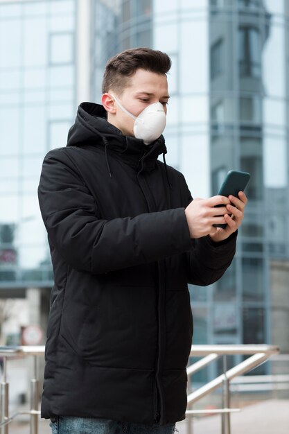Seitenansicht des Mannes mit der medizinischen Maske, die sein Telefon in der Stadt betrachtet