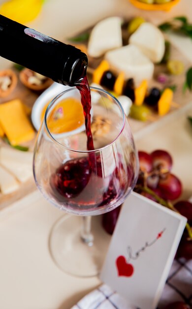 Seitenansicht des Rotweins, der in Glas und verschiedene Arten von Käseoliven-Walnuss-Traube und Liebeskarte auf weißem Hintergrund fließt
