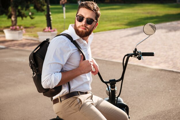 Seitenansicht des ruhigen bärtigen Mannes in der Sonnenbrille mit Rucksack, der draußen auf modernem Motorrad sitzt und wegschaut