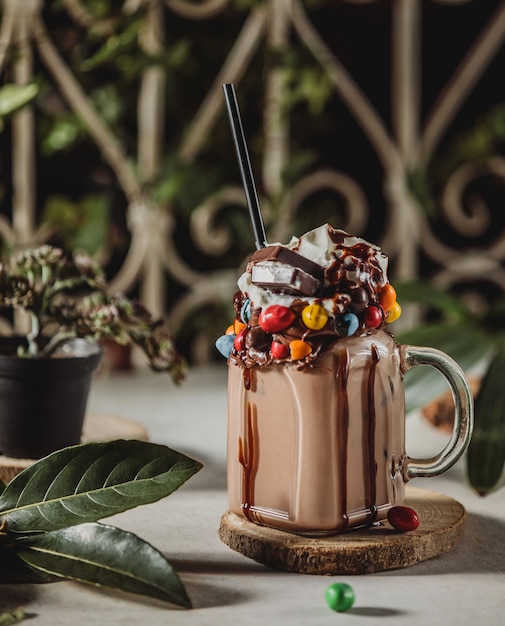 Seitenansicht des Schokoladenmilchshakes mit Schlagsahne, verziert mit Süßigkeiten in einem Glas mit Griff auf einem Holzständer