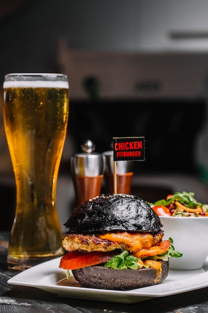 Seitenansicht des schwarzen burgers mit geschmolzenem käse und gemüse des hühnerschnitzels auf einem teller mit einem glas bier auf dem tisch