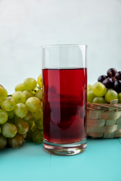 Seitenansicht des schwarzen Traubensaftes im Glas und in den Trauben im Korb und auf blauer Oberfläche und weißem Hintergrund