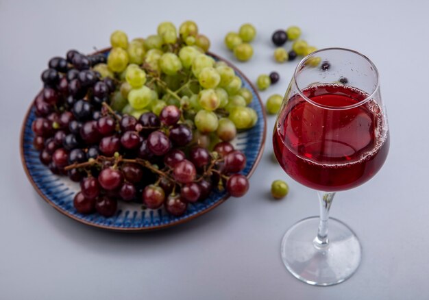 Seitenansicht des schwarzen Traubensaftes im Weinglas und im Traubenteller mit Traubenbeeren auf grauem Hintergrund