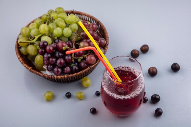 Seitenansicht des schwarzen Traubensaftes mit Trinkröhren im Glas und im Weinkorb mit Traubenbeeren auf grauem Hintergrund