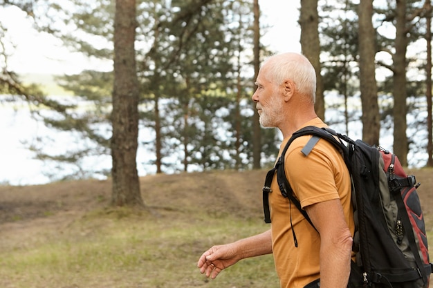 Kostenloses Foto seitenansicht des selbstbestimmten aktiven kahlen männlichen rentners, der rucksack trägt, während er allein in kiefernholz wandert. bärtiger kaukasischer pensionierter mann mit rucksackwanderung entlang der touristischen route im wald