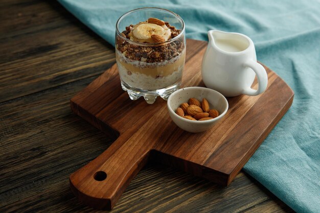 Seitenansicht des Smoothie mit Bananen-Mandel-Walnuss-Milch-Erdnussbutter im Glas und Milch mit Mandeln auf Schneidebrett auf blauem Stoff auf Holzhintergrund