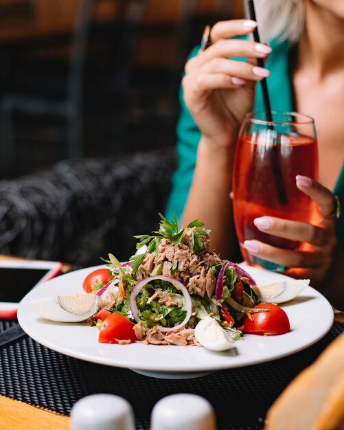 Seitenansicht des Thunfischsalats mit Eiern Zwiebel-Kirschtomaten, die mit frischer Petersilie auf einem weißen Teller mit einer Frau gekrönt werden, die mit Cocktail am Hintergrund sitzt