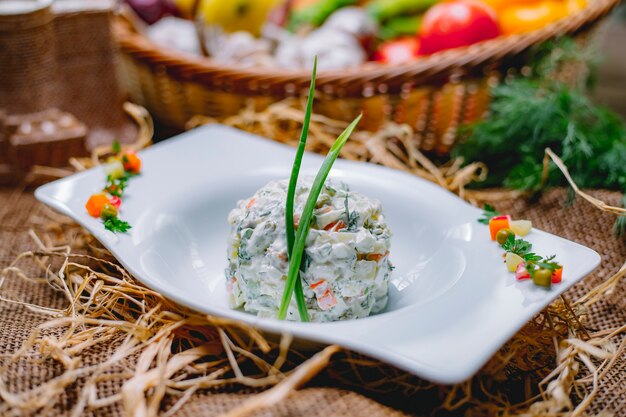 Seitenansicht des traditionellen russischen Oliviersalats mit Huhn verziert mit Frühlingszwiebeln in einer weißen Schüssel auf Strohhintergrund