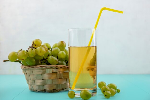 Seitenansicht des Traubensaftes mit Trinkrohr in Glas und Weinkorb mit Traubenbeeren auf blauer Oberfläche und weißem Hintergrund