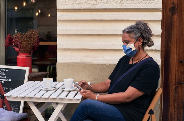 Seitenansicht einer Frau mit einer Gesichtsmaske, während sie ihr Telefon auf dem Tisch im Freien eines Cafés benutzt