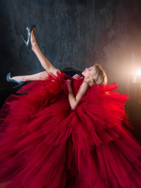 Seitenansicht einer fröhlichen Blondine in einem üppigen roten Kleid sitzt auf einem Stuhl und hebt ihre Beine