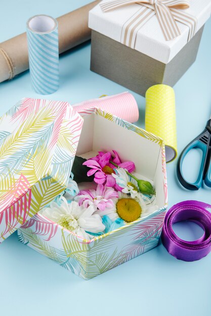 Seitenansicht einer Geschenkbox gefüllt mit bunten Chrysanthemenblumen mit Gänseblümchen- und Scherenrollen Papier und lila Band auf blauem Hintergrund
