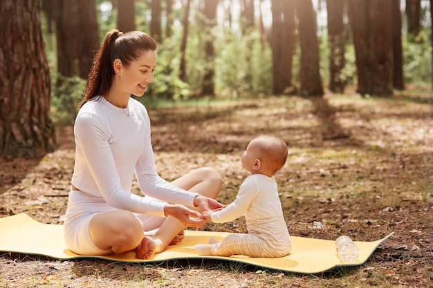 Seitenansicht einer glücklichen jungen sportlichen mutter, die mit ihrem säugling auf karemat im wald sitzt