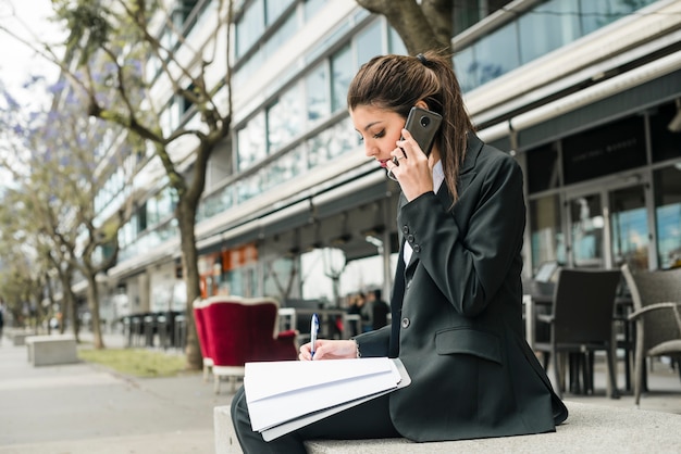 Seitenansicht einer jungen Geschäftsfrau, die außerhalb des Gebäudeschreibens auf Ordner mit Stift sitzt