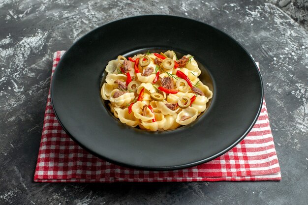 Seitenansicht einer köstlichen Conchiglie mit Gemüse und Grün auf einem Teller und Messer auf rotem, abgestreiftem Handtuch auf grauem Hintergrund