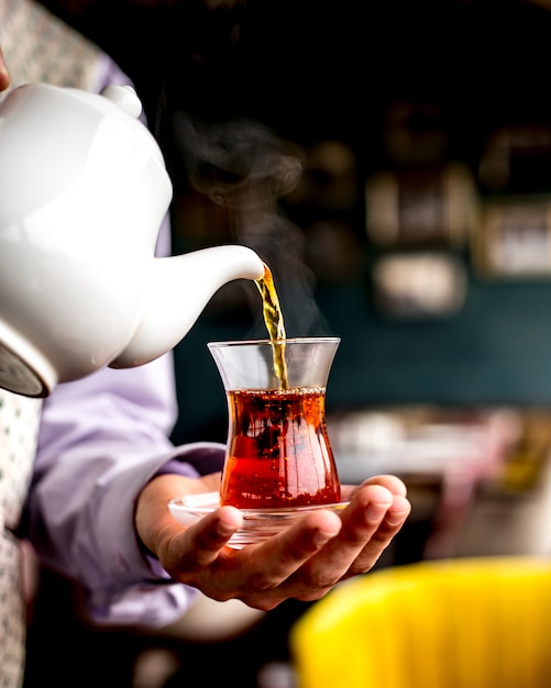 Seitenansicht einer Person, die schwarzen Tee aus weißer Keramik-Teekanne in Armudu-Glas gießt
