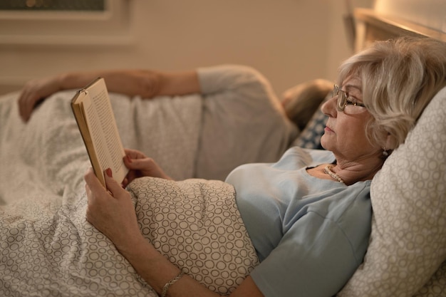 Seitenansicht einer reifen Frau, die sich im Bett hinlegt und ein Buch liest Ihr Mann schläft im Hintergrund