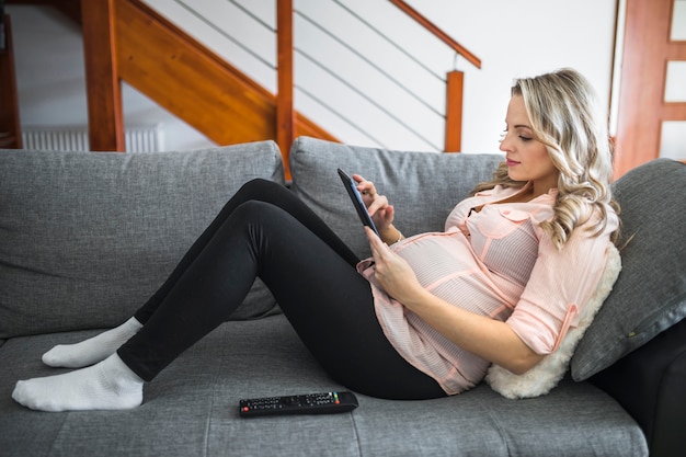 Seitenansicht einer schönen schwangeren Frau, die auf Bett unter Verwendung der digitalen Tablette liegt