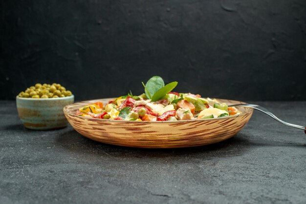 Seitenansicht einer Schüssel Gemüsesalat mit Gabel und Erbsen auf der Seite auf dunkelgrauem Tisch auf dunkelgrauem Hintergrund