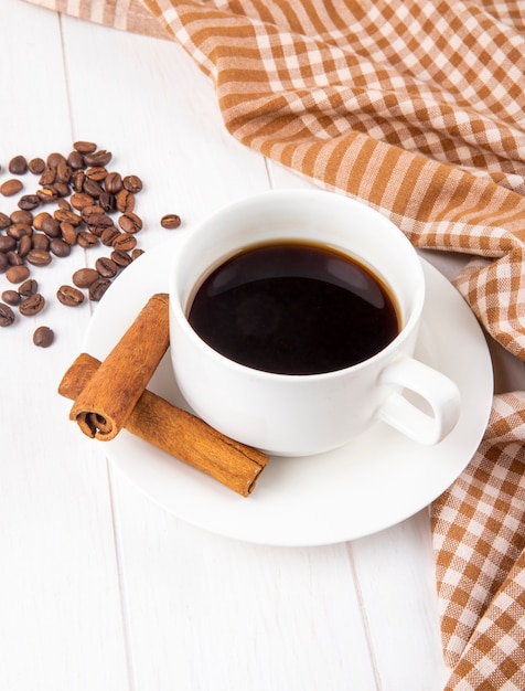 Seitenansicht einer Tasse Kaffee mit Zimtstangen und Kaffeebohnen verstreut auf weißem hölzernem Hintergrund