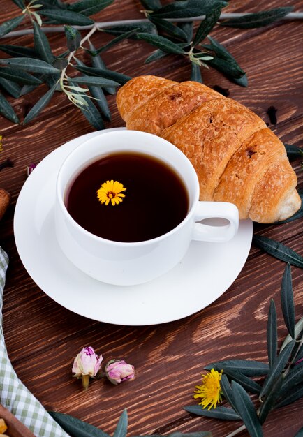 Seitenansicht einer Tasse Tee mit Croissant und Löwenzahn auf Holz