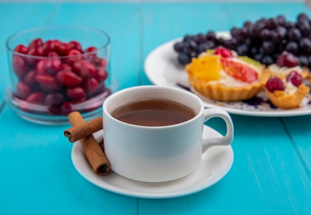 Seitenansicht einer Tasse Tee mit Zimtstangen mit Kornelkirschenbeeren auf einer Glasschale auf einem blauen hölzernen Hintergrund