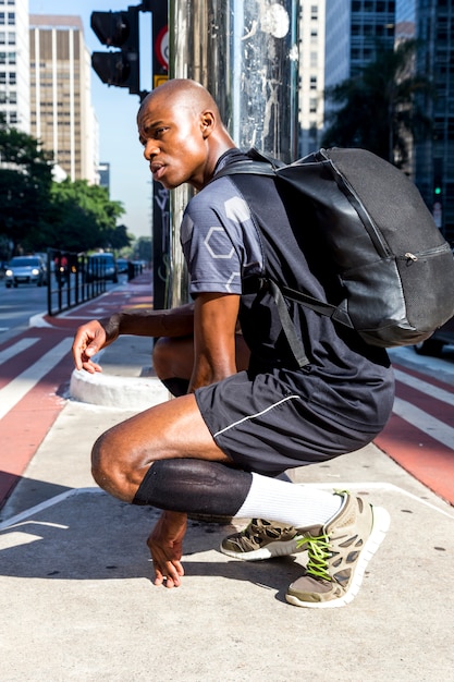 Seitenansicht eines afrikanischen jungen sportlichen Mannes mit seinem Rucksack, der mitten in Straße sich duckt