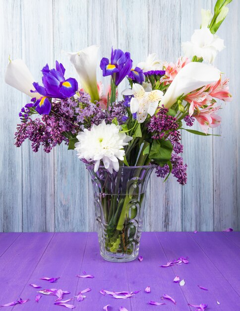 Seitenansicht eines Blumenstraußes der weißen Farbe Calla-Lilien mit dunkelvioletten Iris-lila weißen Gladiolen und rosa Alstroemeria-Blumen in einer Glasvase auf lila Oberfläche auf grauem Holzhintergrund