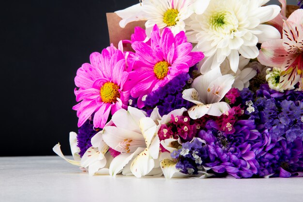 Seitenansicht eines Blumenstraußes von rosa weißen und lila Farbstatice-Alstroemeria- und Chrysanthemenblumen in Bastelpapier, das auf weißer Oberfläche bei schwarzem Hintergrund liegt