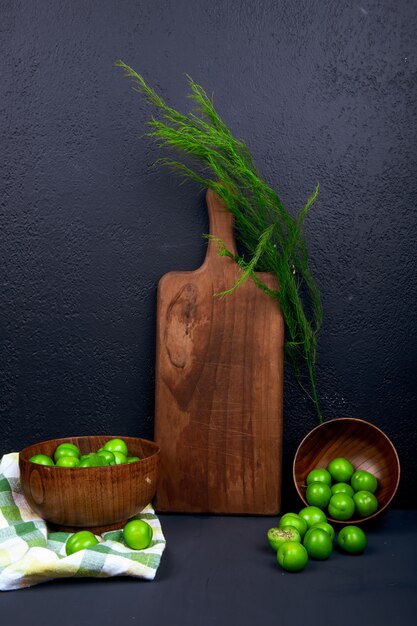 Seitenansicht eines hölzernen Schneidebretts mit Fenchel und sauergrünen Pflaumen in Holzschalen auf schwarzem Tisch