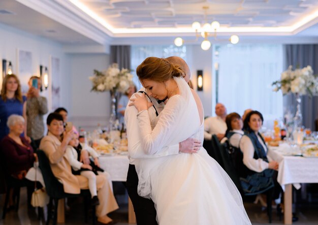 Seitenansicht eines hübschen Ehepaars, das sich umarmt und den ersten Hochzeitstanz in einem gemütlichen Restaurant tanzt, während ihre Familie und Gäste dabei sind