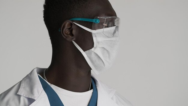 Seitenansicht eines jungen afroamerikanischen männlichen Arztes mit Schutzbrille und medizinischer Maske, der selbstbewusst vor weißem Hintergrund aussieht