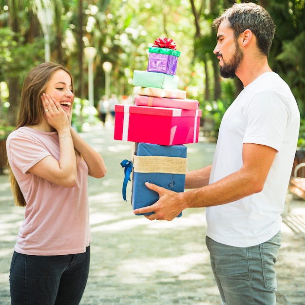 Seitenansicht eines jungen Mannes, der seiner überraschten Freundin Stapel Geschenke gibt