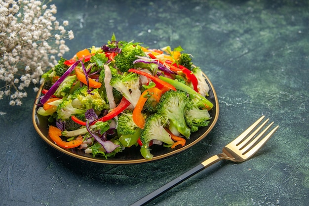 Seitenansicht eines köstlichen veganen Salats in einem Teller mit verschiedenem Gemüse und weißer Gabelblume auf dunklem Hintergrund