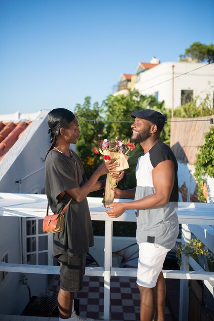 Seitenansicht eines romantischen afrikanischen schwulen Paares mit Blumen. Zwei lächelnde Männer, die auf dem Dach stehen und sich ansehen, ein Mann, der einem anderen Mann schöne Blumen gibt. LGBT-Paare lieben, Lifestyle-Konzept