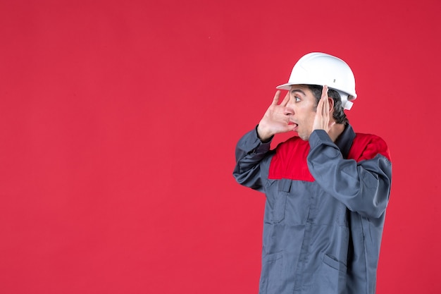 Seitenansicht eines schockierten jungen Architekten in Uniform mit Schutzhelm auf isolierter roter Wand