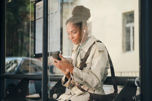 Seitenansicht eines schönen, lässigen afroamerikanischen Mädchens im stilvollen Trenchcoat mit Kaffee, das glücklich mit dem Handy an der Bushaltestelle telefoniert