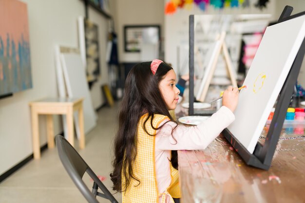 Seitenansicht eines schönen Mädchens während eines Kunstunterrichts. Hispanisches Kind, das vor einer Leinwand auf einer Staffelei sitzt und lernt, wie man malt