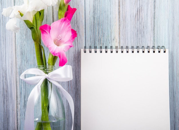 Kostenloses Foto seitenansicht eines skizzenbuchs mit weißen und rosa farbe gladiolenblumen in einer glasflasche auf grauem hölzernem hintergrund