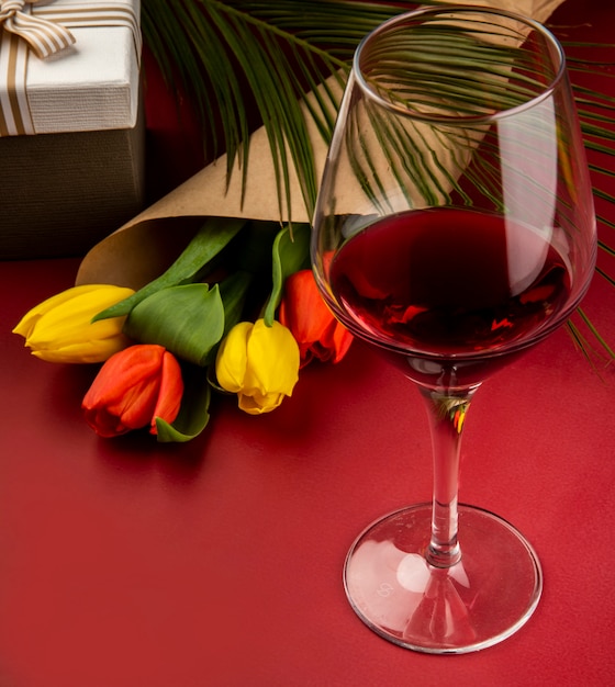 Seitenansicht eines Straußes roter und gelber Farbtulpen in Bastelpapier und eines Glases Wein auf rotem Tisch