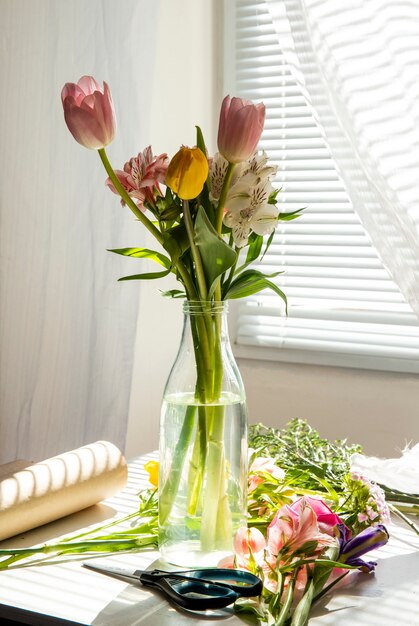 Seitenansicht eines Straußes von rosa und gelben Farbtulpen mit Alstroemeria-Blumen in einer Glasflasche auf dem Tisch