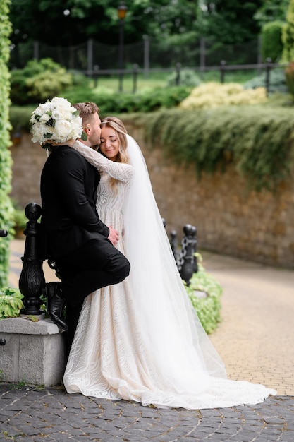 Seitenansicht eines süßen Mädchens in Spitzenkleid mit Kleiderschleppe, das ihren Bräutigam umarmt und die Augen während des Hochzeitsspaziergangs im Park schließt